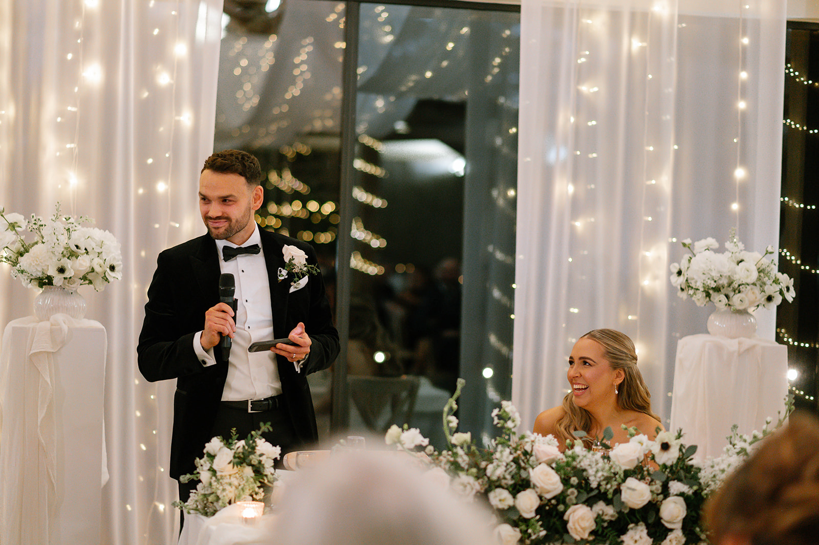 Winter wedding at a glass-walled barn at dusk, candlelit interior glowing through snowy trees